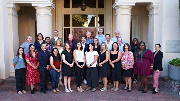 A group of smiling people posing outside Nobili Hall on the Santa Clara campus.