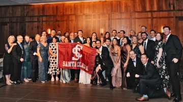 A group of smiling people in wedding outfits, including a bride and a groom, holding a red 