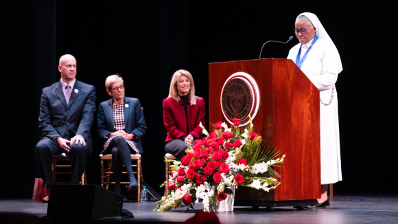 A woman in a white gown and wimple, speaking at a podium bearing the Santa Clara University seal.