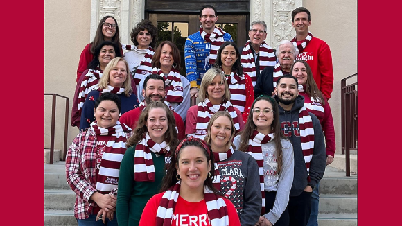 19 smiling people in holiday themed outfits outside Bannan Alumni House.