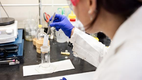 A person in safety goggles dips a test paper into a beaker