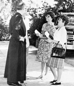 A priest talks to two women holding books outside.