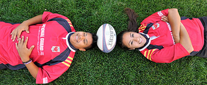 Two women in red jerseys lying on grass with a rugby ball.