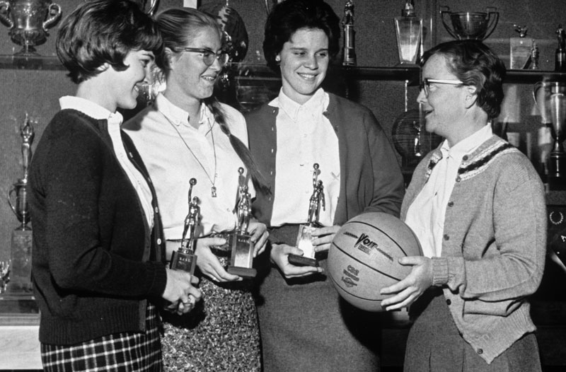 Four women holding basketball trophies, one holding a basketball, in a 1964 photograph.
