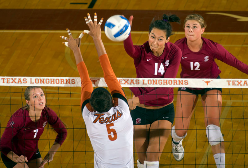 Three volleyball players in red jerseys blocking a spike from an opponent.