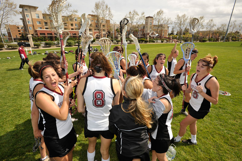 A women's lacrosse team huddles on a grassy field.