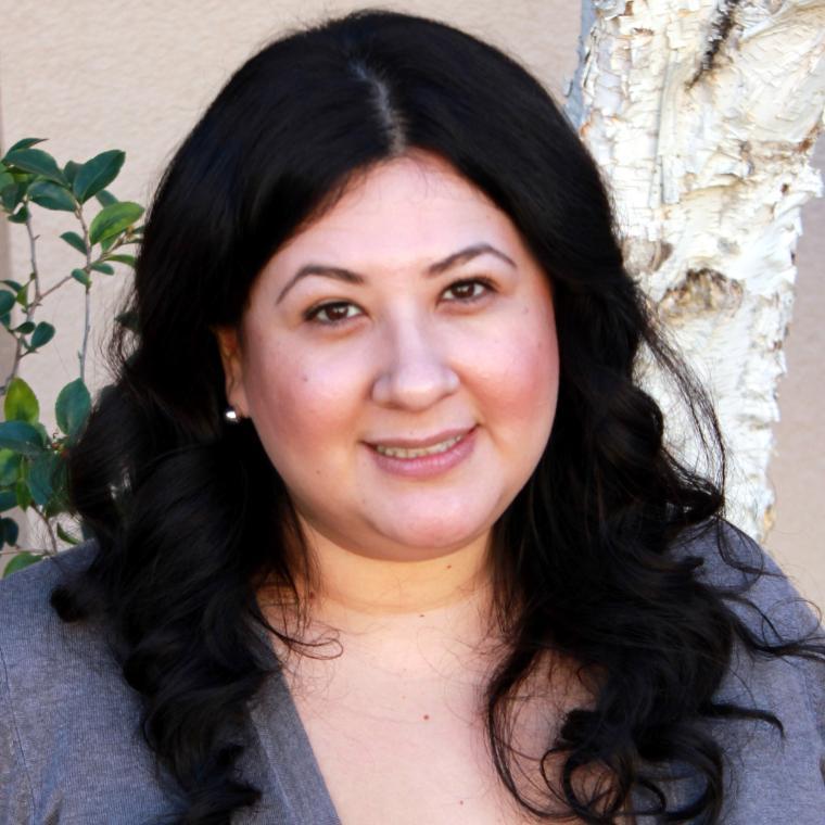 A person smiling in front of a beige background with greenery.