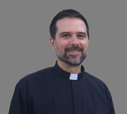 Man in clerical attire smiling against a grey background.