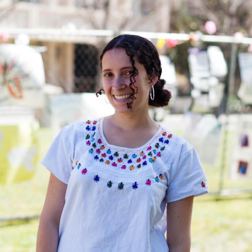 Woman smiling, standing outdoors with trees and structures in the background.