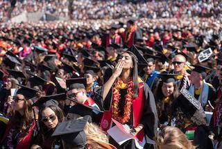 A couple wearing graduation gowns kiss in a large crowd.