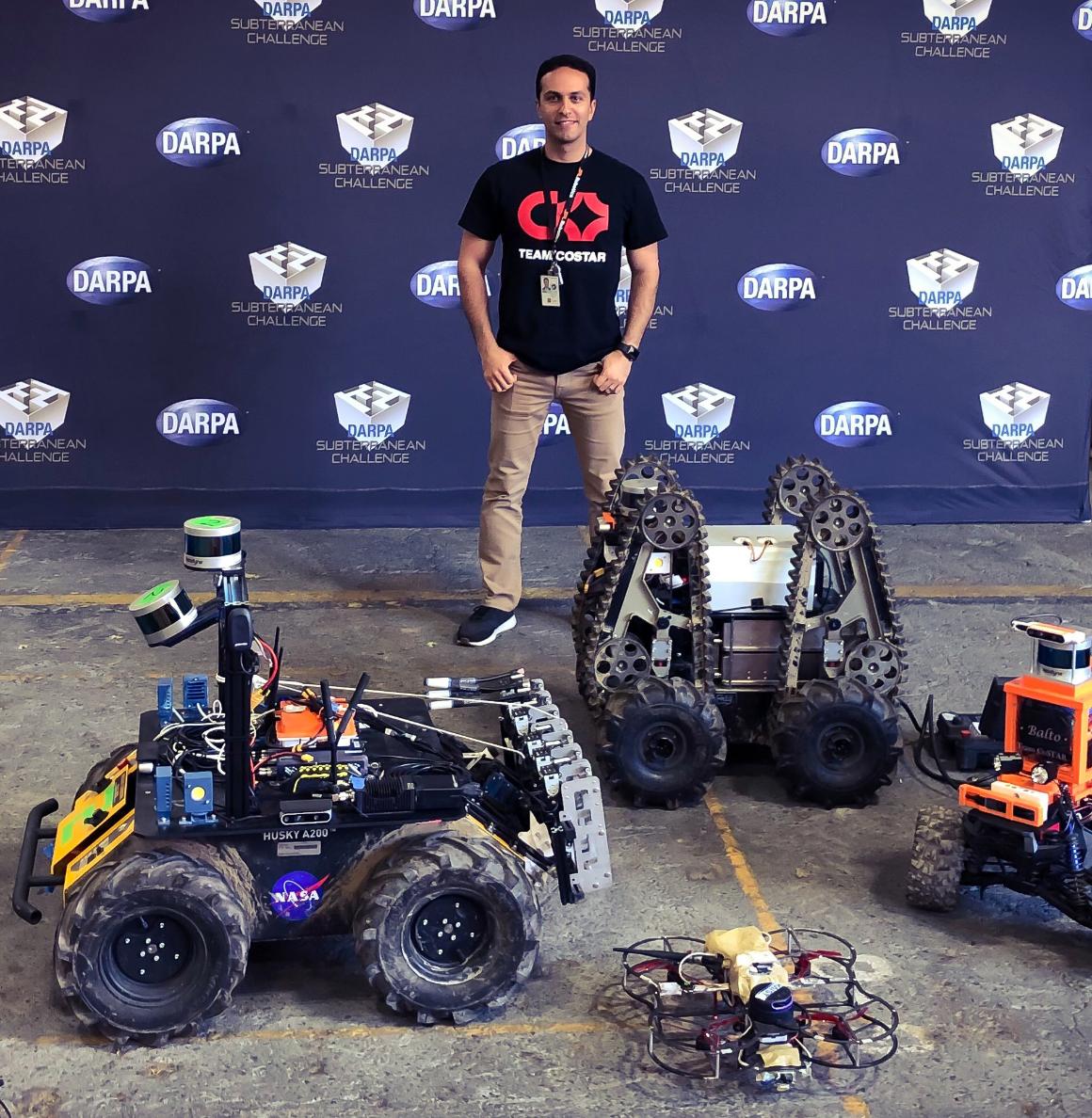 Kamak Ebadi standing in front of a DARPA sign with a variety of robots at his feet