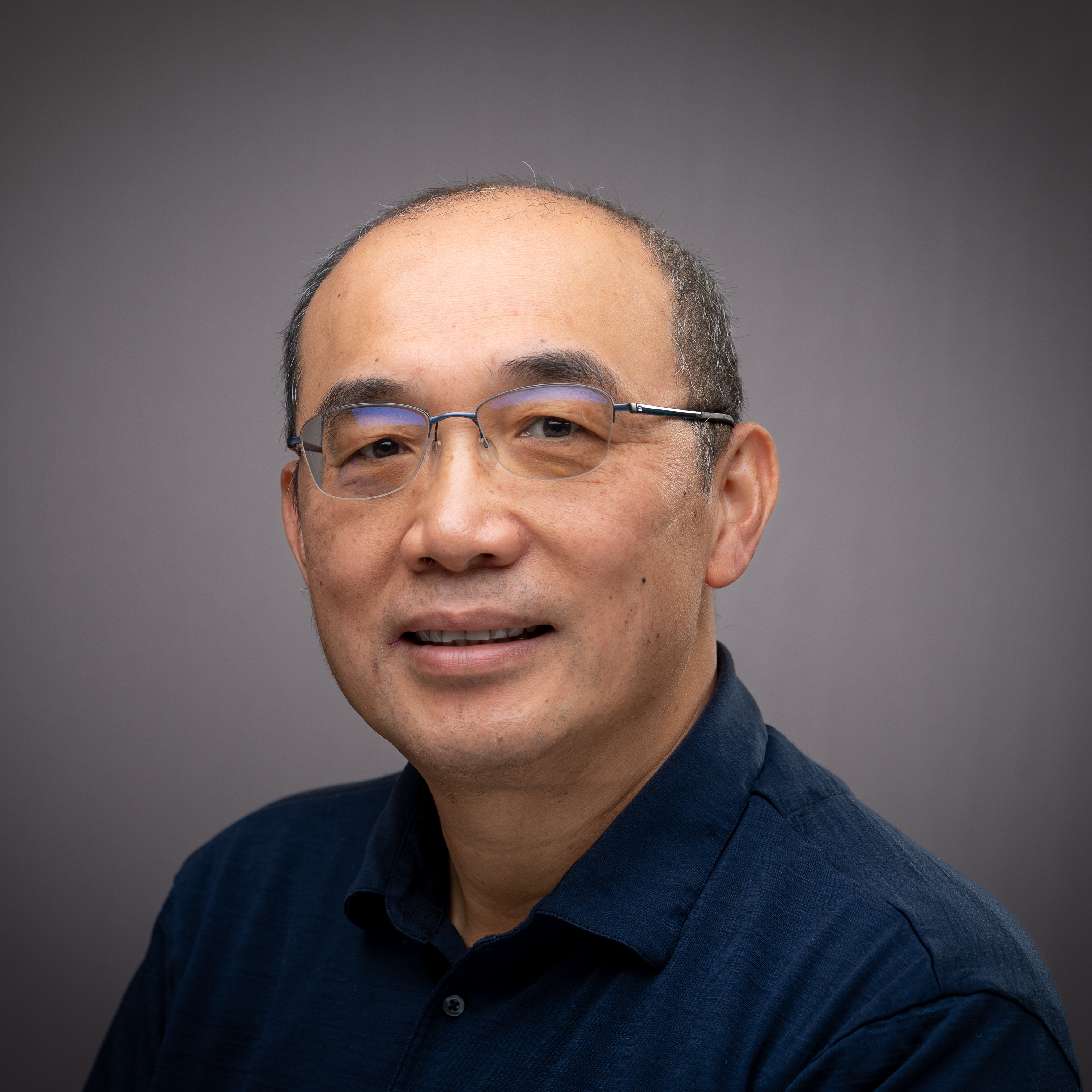 A man wearing glasses and a dark shirt, smiling against a gray background.