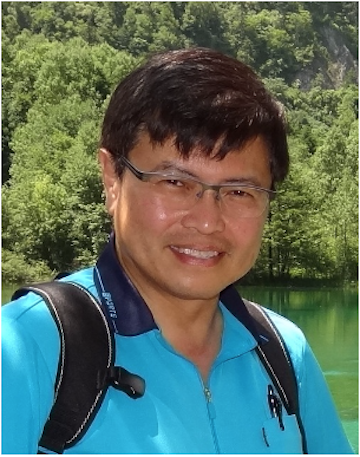 A man smiling outdoors with greenery in the background.