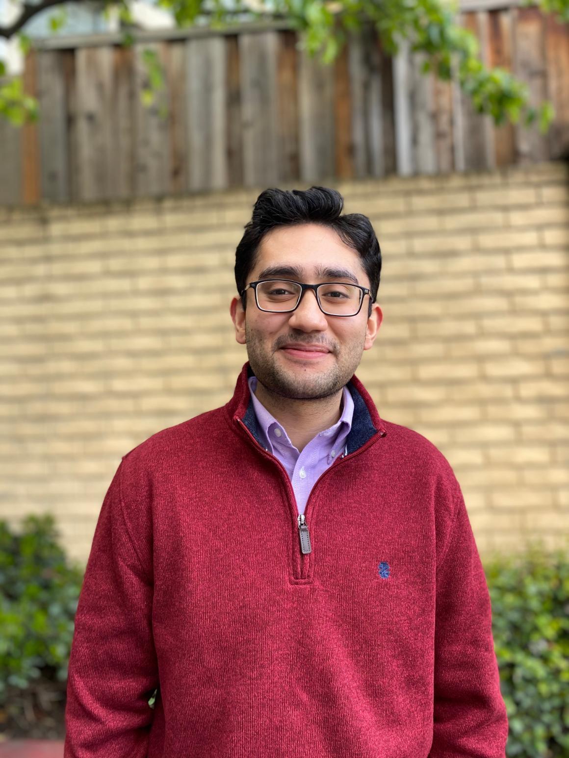 A person smiling, wearing a red jacket, stands outdoors near a brick wall.