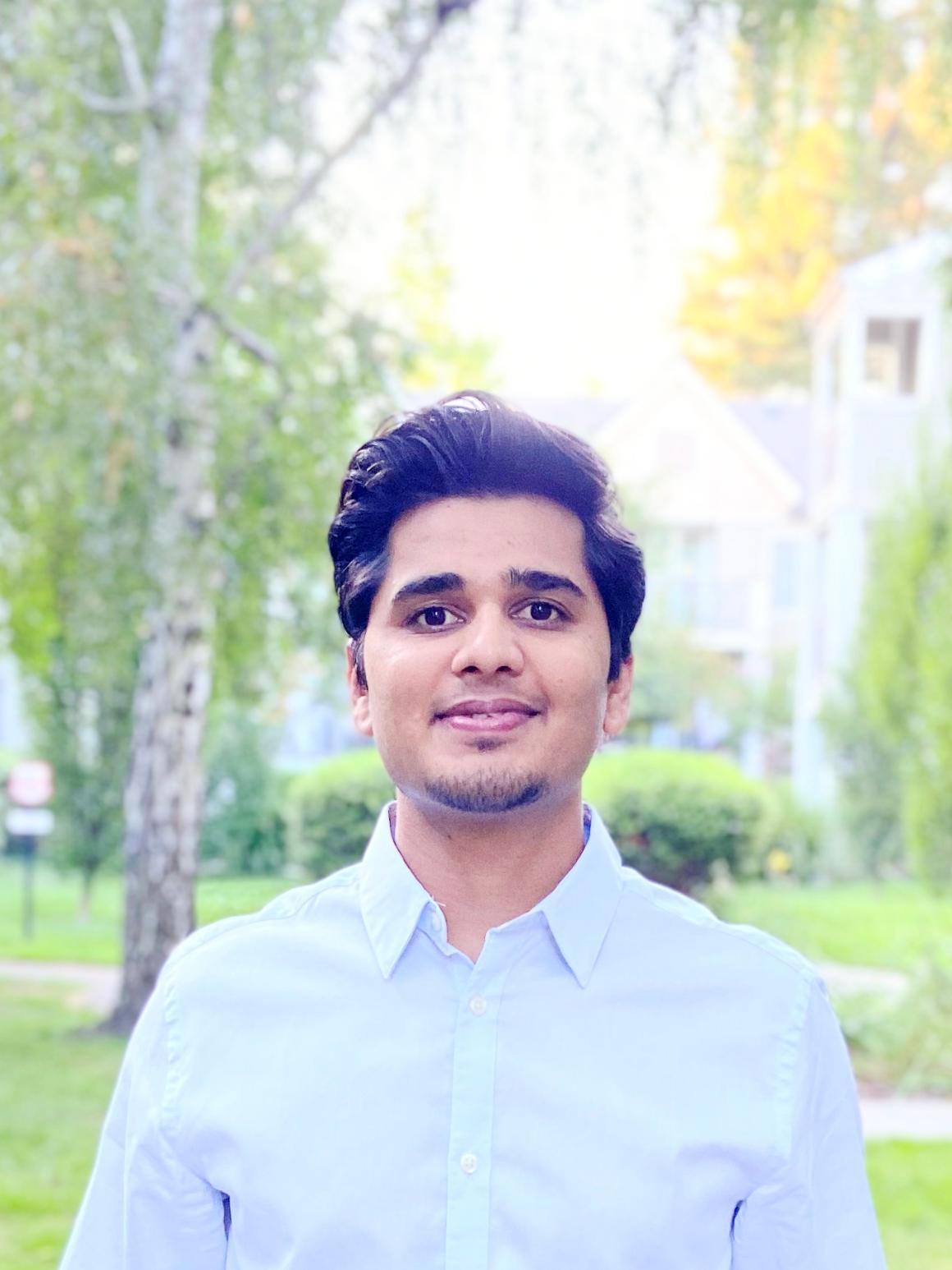 A person in a white shirt standing outdoors, greenery in the background.