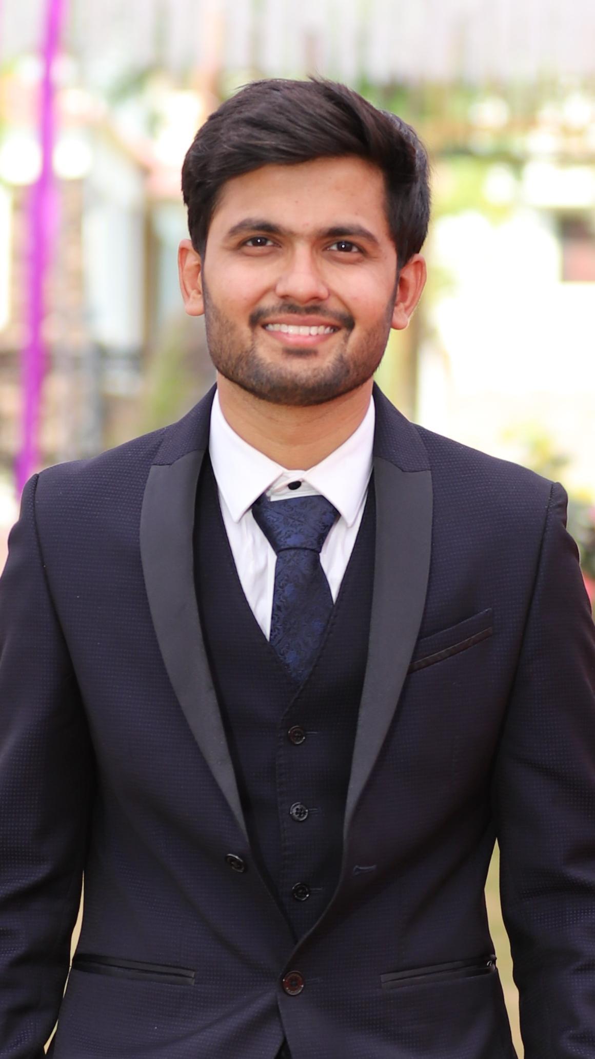 A person in a dark formal suit, standing and smiling.