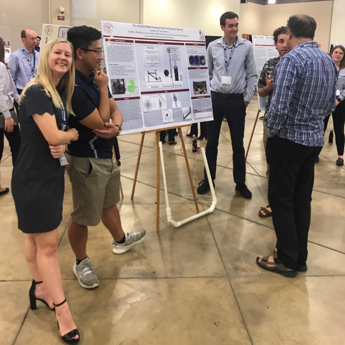 Female student smiles at the camera while male teammate talks about their poster with visitors