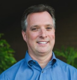 A person in a blue shirt smiling against a dark background with green leaves.