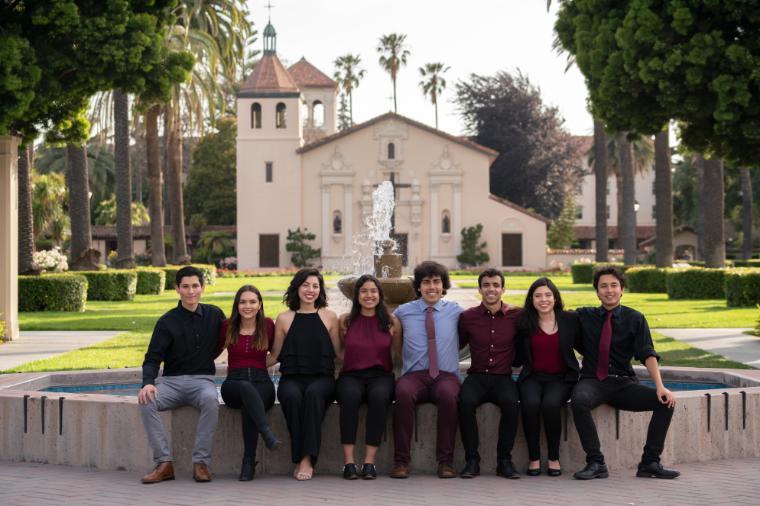 Group of people in formal attire seated outside a historic building on a sunny day.