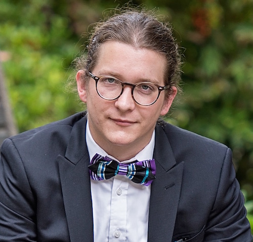 Man in a suit with a bow tie and glasses, outdoor background.