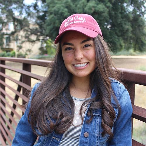 A person in a denim jacket and red cap smiling on a bridge.