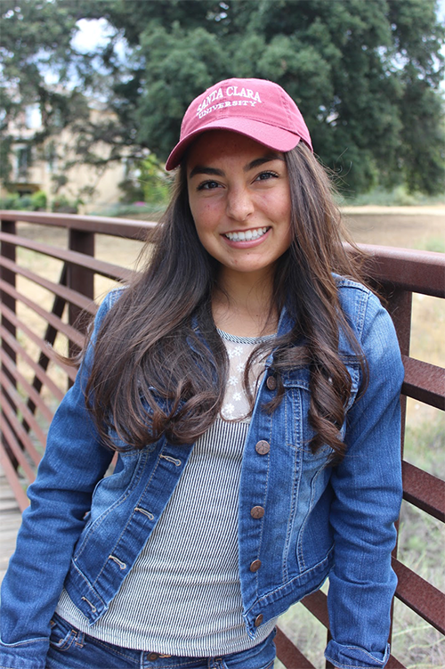 A person in a denim jacket and red cap smiling on a bridge.