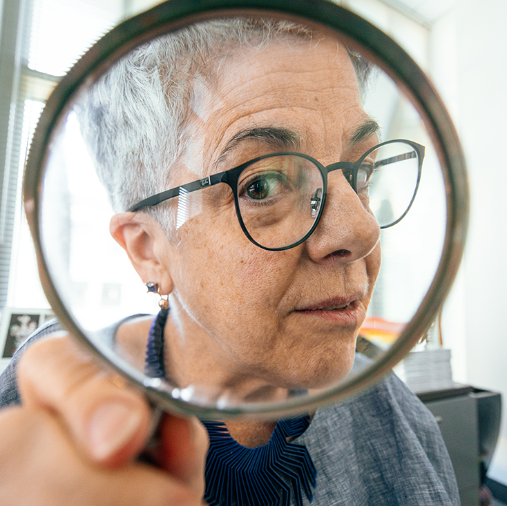 Andrea Pappas in an office with bookshelves peering through a magnifying glass at the camera