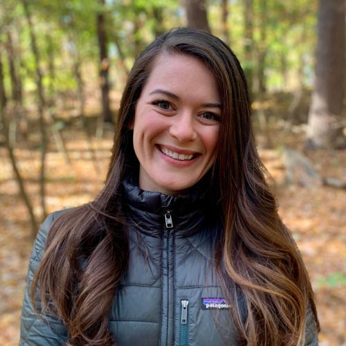 A person smiling wearing a dark jacket in a forest setting.