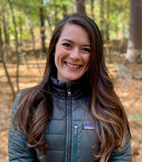 A person smiling wearing a dark jacket in a forest setting.