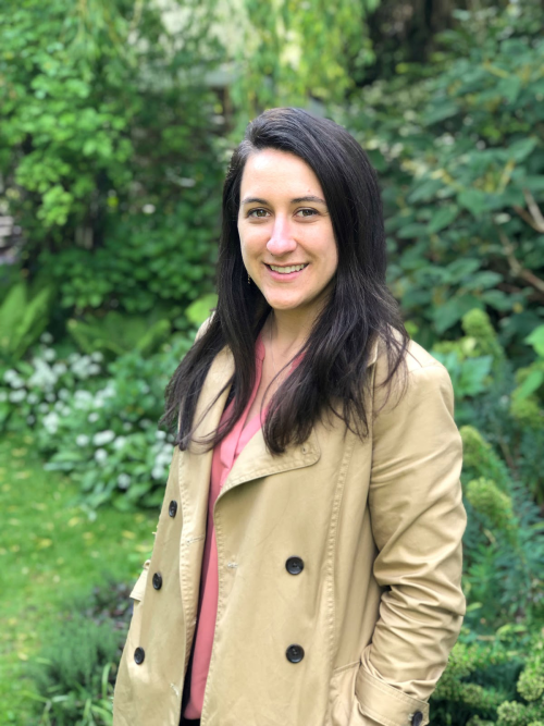 A person in a beige coat smiling in a garden.