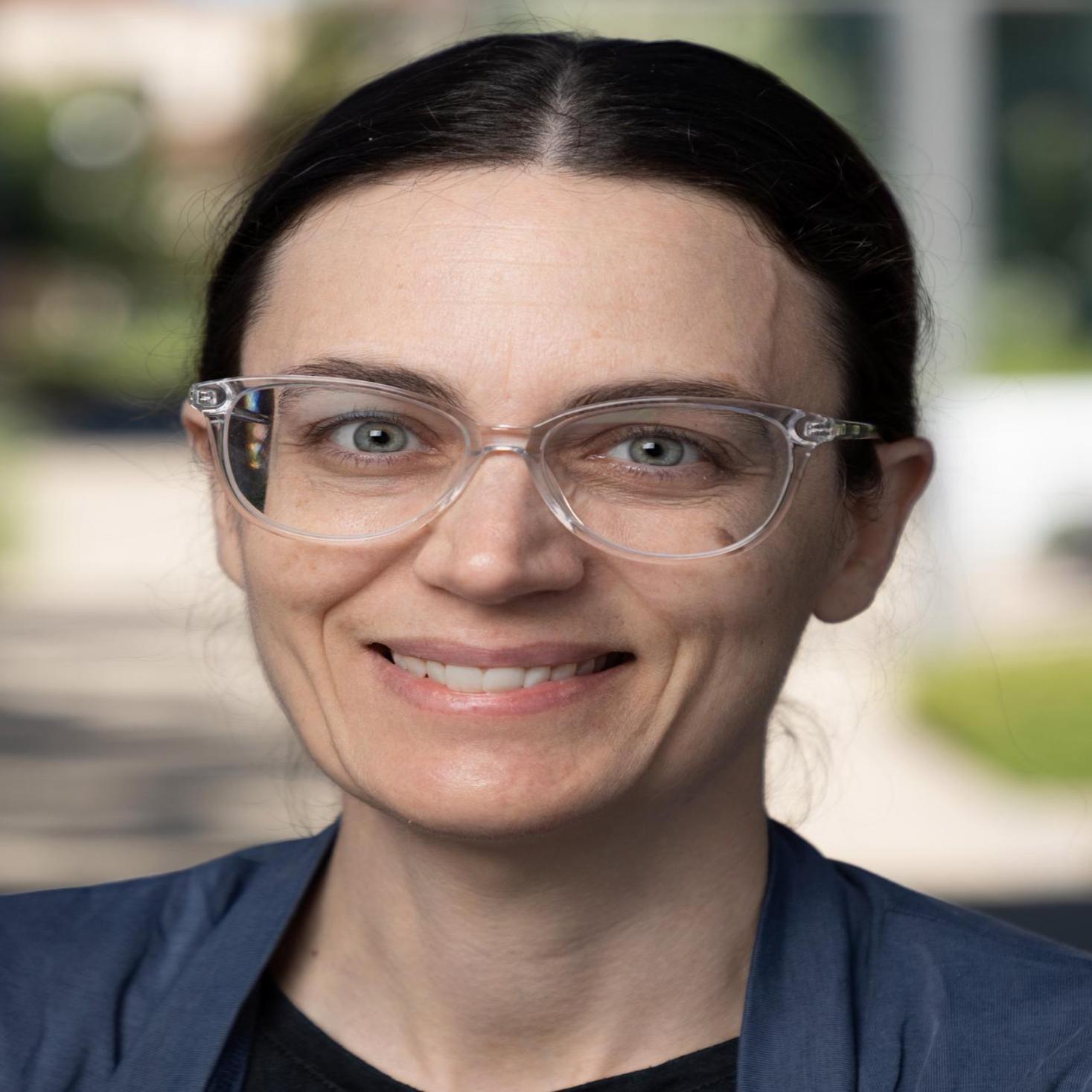 A person wearing glasses and a blue cardigan, smiling outdoors.