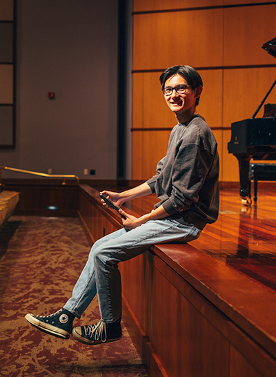 Male student sitting on the edge of a stage with a grand piano in the background