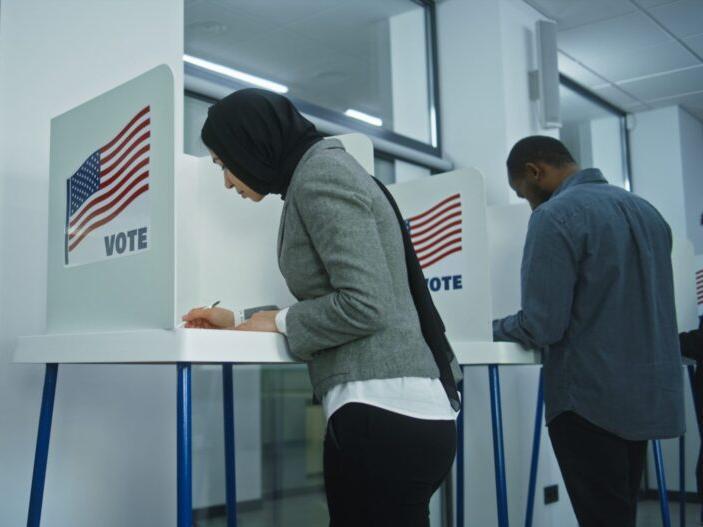 Muslim woman in grey jacket and black hijab and pants in a voiting booth