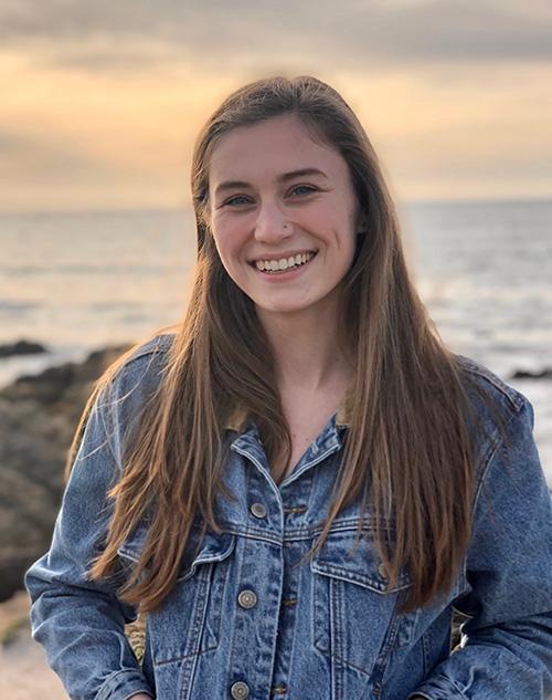 A person smiling with a sunset over the ocean in the background.