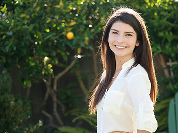 A woman smiling outdoors in front of green foliage.