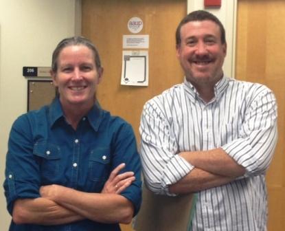 Two people standing with arms crossed, smiling, in front of a door. image link to story