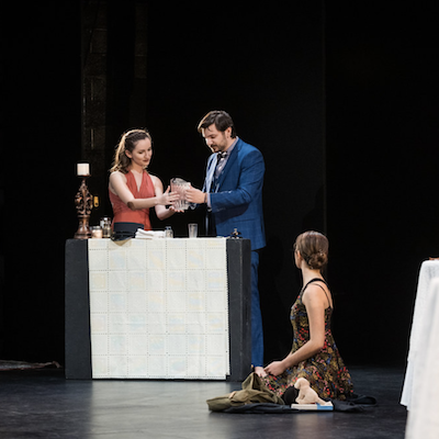 Four people performing a scene on stage with a table and props.