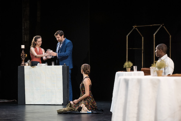 Four people performing a scene on stage with a table and props.