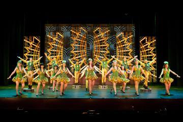 Male and female tap dancers wearing gold and green costumes in the big 42nd Street dance number set against a backdrop of lightbulbs in fan shapes