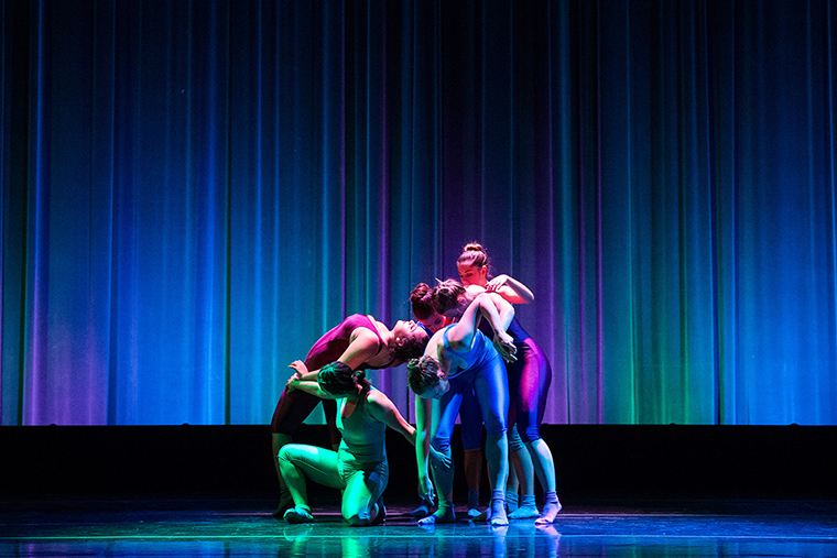 Dancers perform onstage in colorful costumes against a blue-lit curtain backdrop.