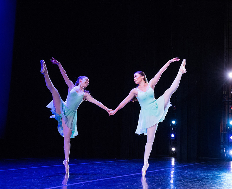 Two dancers performing synchronized ballet moves on stage, holding hands.