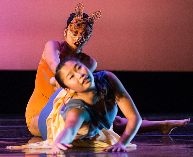 Two dancers performing expressive choreography on stage with a colorful background.