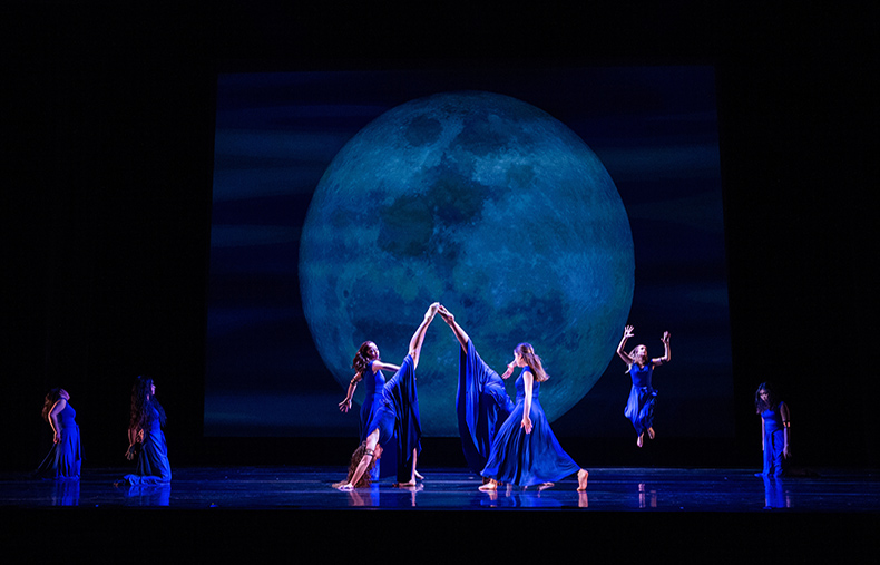 Performers in blue costumes dance under a large moonlit backdrop.