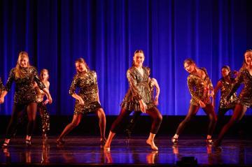 8 female jazz dancers in black and gold costumes on a stage with a bright blue backdrop