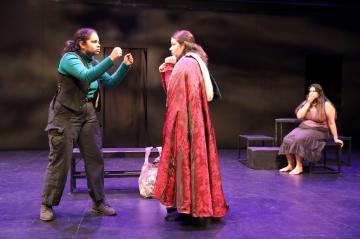 A woman in black and green with her fists up facing a woman in a red velvet cloak with a fur trimmed hood whose bag is on the ground