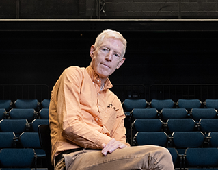 Brian Thorstenson sitting in Fess Parker studio Theatre wearing an orange shirt, khaki pants and Converse boots