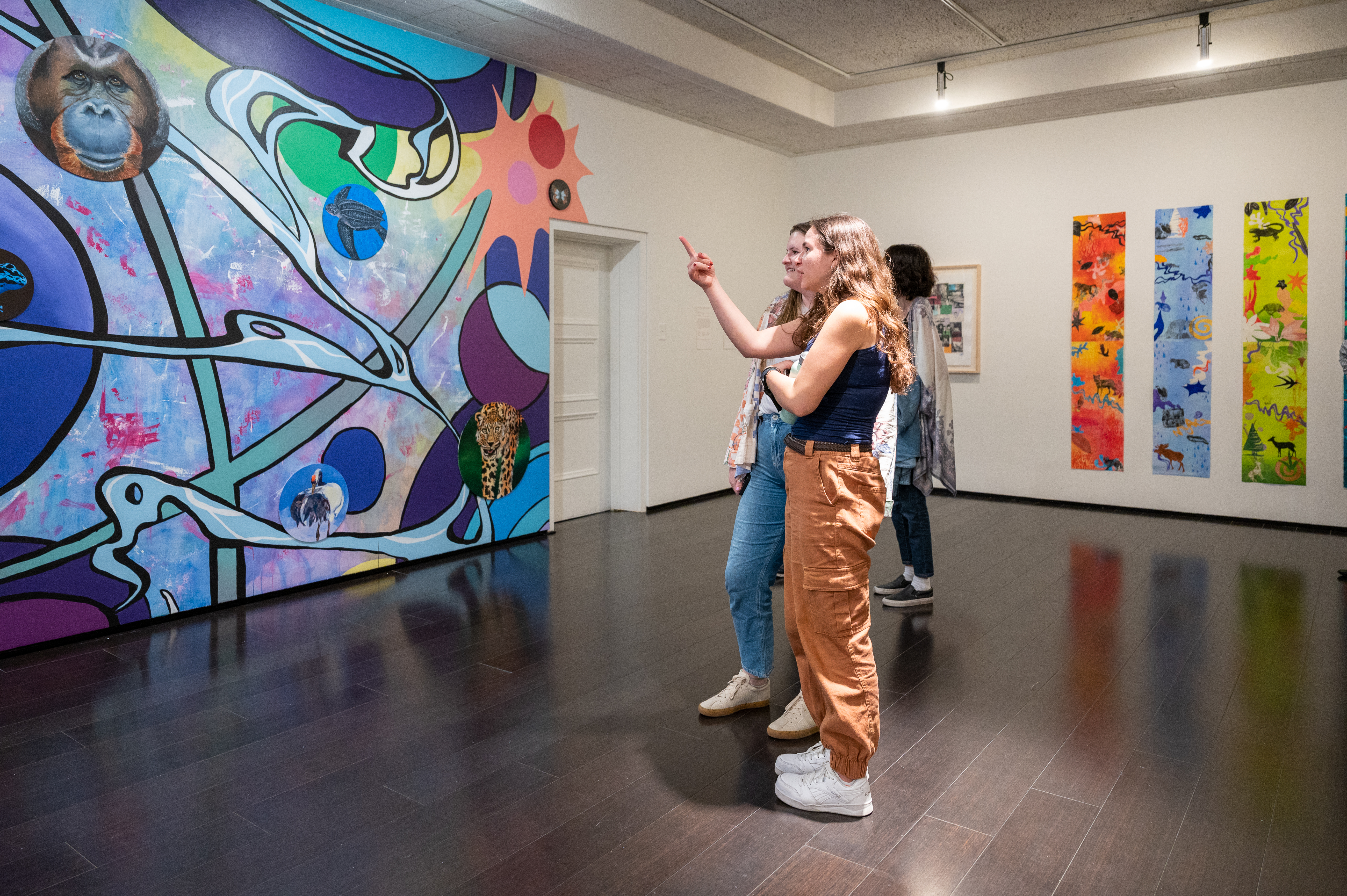 Students in gallery pointing to aspects of large mural with prints hanging in the backgroud