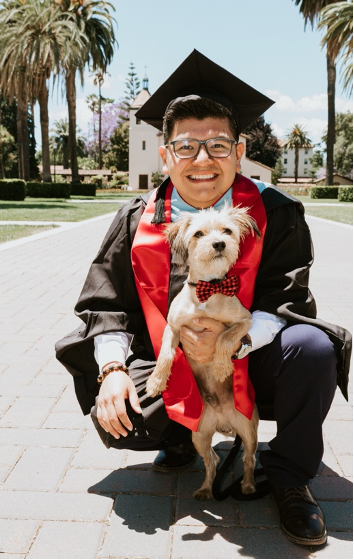 Graduate in cap and gown holding a small dog on a sunny day.