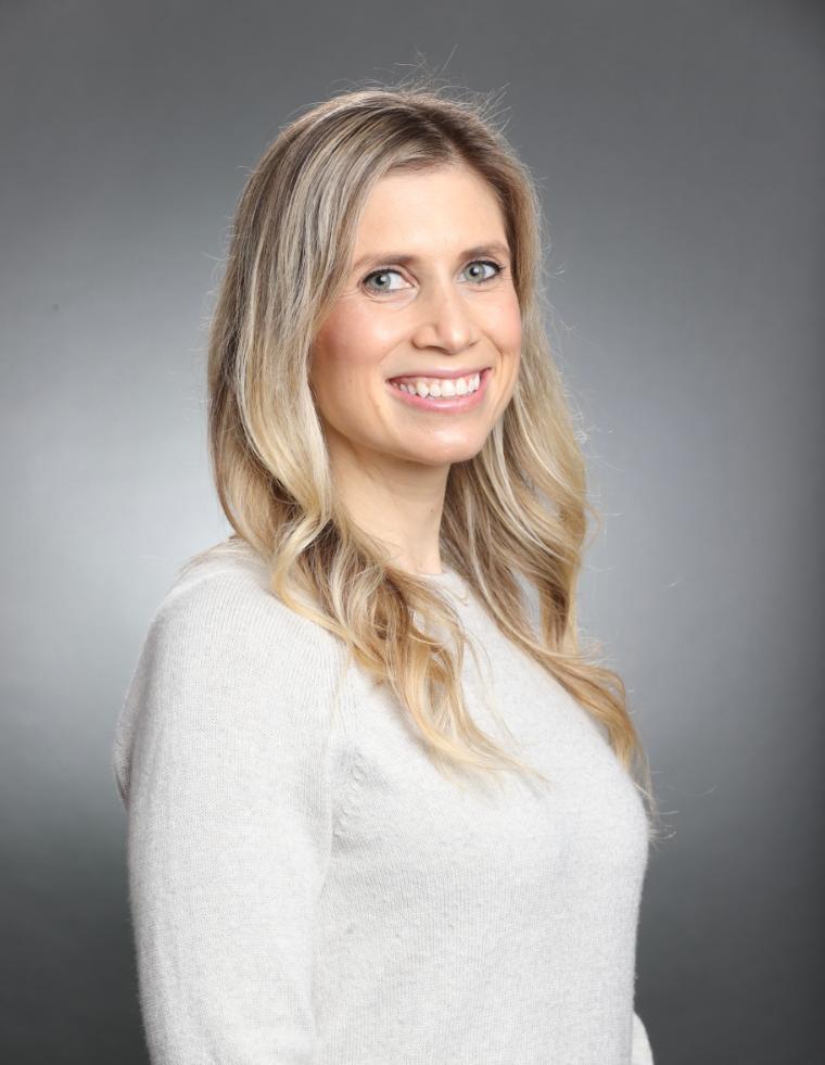 A woman smiling, wearing a white shirt against a gray background.