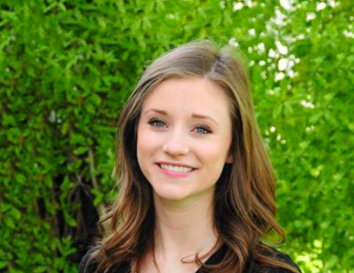 A person smiling outdoors with green foliage in the background.